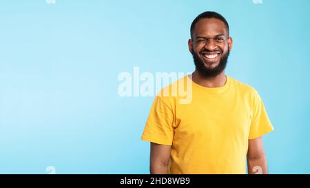Glücklicher bärtiger junger schwarzer Mann in gelbem T-Shirt, der die Kamera anschaut und auf blauem Hintergrund zwinkert, Banner mit Kopierraum Stockfoto