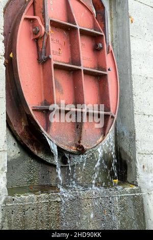 Rundes rundes rotes Schleusentor mit Wasser, das unten ausströmt. Auf Betonwand montiert. Stockfoto