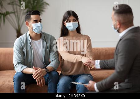 Glückliches Paar in Gesichtsmasken, das im Büro mit dem Therapeuten die Hände schüttelt Stockfoto