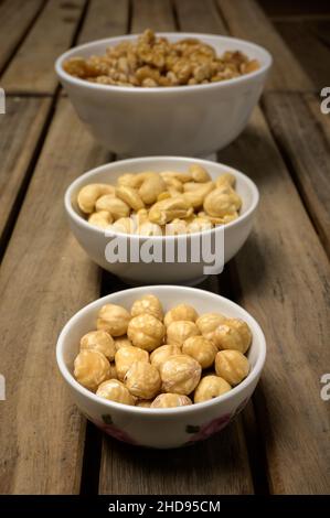 Haselnüsse, Cashewnüsse und Walnüsse in verschiedenen Gläsern auf einem Holztisch in der Draufsicht und in dunkler Umgebung. Stockfoto