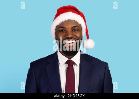Lächelnder afroamerikanischer Geschäftsmann in weihnachtsmütze, der auf blauem Hintergrund posiert Stockfoto