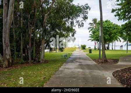 Airlie Beach, Queensland, Australien - 2022. Januar: Menschen, die entlang eines Fußweges am Strand spazieren, mit umgestuften Einkaufsbuden neben einem kleinen Wald Stockfoto