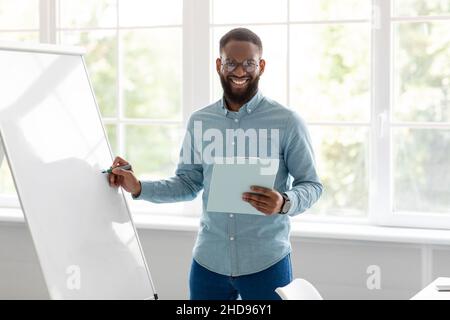 Fröhlich hübsch junge afroamerikanische männliche Tutor, Lehrer in der Nähe von Tafel schaut auf Kamera im Home Office-Interieur Stockfoto