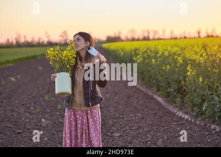 Eine verträumte Frau zieht bei Sonnenuntergang ihre Schutzmaske in einem Frühlingsrappesaatfeld aus. Fröhliche Emotionen. Quarantäne- und Covid-Virenbeendigungskonzept. Stockfoto