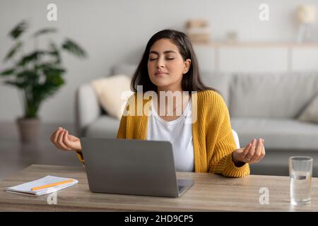 Stressmanagement am Arbeitsplatz. Ruhige arabische Frau, die mit geschlossenen Augen vor dem Laptop meditiert und am Schreibtisch sitzt Stockfoto