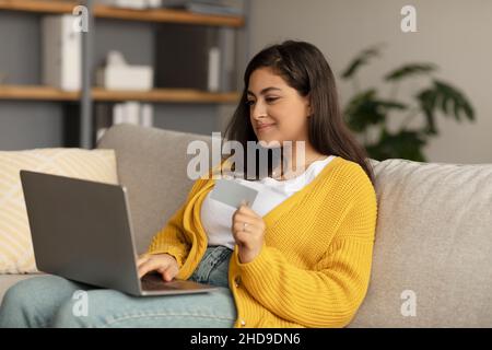 Junge Dame aus dem Nahen Osten mit Kreditkarte und Laptop, sitzt auf der Couch, kauft online und bezahlt kontaktlos Stockfoto