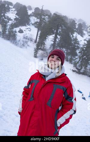 Portrait eines glücklichen Teenagers, das lächelt und im Winter in warmen roten Mantel bei Schnee gekleidet ist Stockfoto