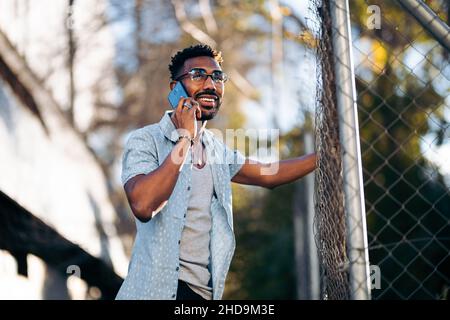 Ein hübscher Afro-Mann, der sein Smartphone auf der Straße benutzte und lächelte, an einem Zaun gelehnt Stockfoto