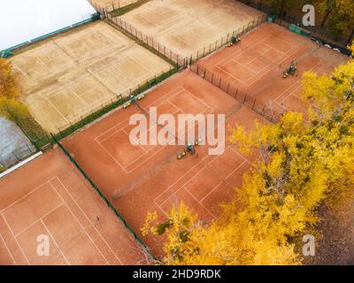 Fliegen Sie über die orangefarbenen Sandtennisplätze im Stadtpark. Leuchtend gelbe Herbstbäume in der Nähe des Sportrekreationsgebiets. Luftaufnahme von Baumkronen auf buntem Kharkiv, trav Stockfoto