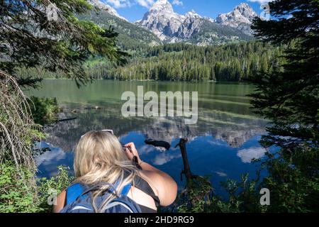 Die blonde Wandererin kniet sich nieder, um den Taggart Lake im Grand Teton Natinoal Park zu fotografieren Stockfoto