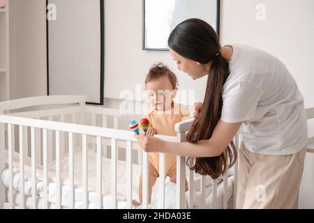 Junge Mutter gibt ihrem kleinen Baby Spielzeug in der Krippe zu Hause Stockfoto