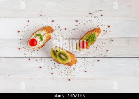 Drei Mini-Toasts umgeben von roten Beeren in Herzform, auf weißem Holzhintergrund, Überraschung zum Valentinstag, Draufsicht Stockfoto