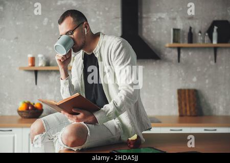 Porträt eines Mannes, der Kaffee trinkt und eine Brille trägt und ein Buch liest, während er sich in der Küche ausruht. Kaffeepause. Stockfoto