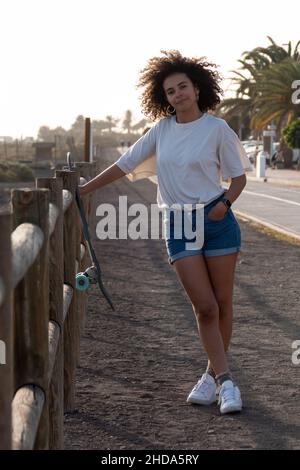 Skater junge Frau mit Afro-Frisur auf einem Zaun gelehnt. Sie lächelt und trägt Sommerkleidung. Stockfoto