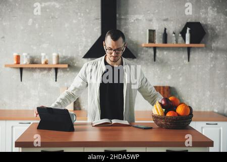 Vorderansicht eines Geschäftsmanns, der zu Hause in der Küche arbeitet. Intelligentes Arbeiten mit Tablets und Notebooks während der covid Pandemie. Stockfoto