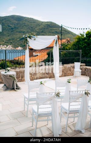 Weiße Stühle mit Blumensträußen und Stoffen stehen vor einem Hochzeitbogen in einem Innenhof mit Blick auf das Meer Stockfoto