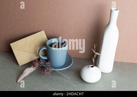 Stillleben von Kaffeetasse und Umschlag und Keramikvase mit trockener Pflanze. Grauer Hintergrund Stockfoto