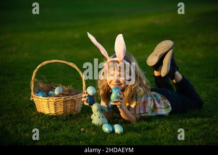 Kinder jagen ostereier. Kleiner Junge, der auf dem Gras liegt und Eier findet. Kind mit ostereiern und Hasenohren, Außenportrait. Kleiner Hase Junge Stockfoto