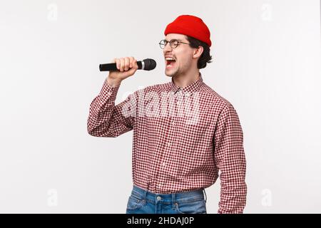 Unbeschwert aufgeregter und witziger kaukasischer bärtiger Mann in roter Mütze und kariertem Hemd, Augen schließen als passioniert gesangtes Karaoke-Lieblingslied, halten Stockfoto