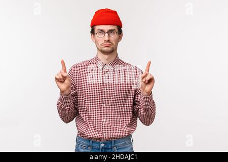 Taillenhöhe Hochformat verärgert und enttäuscht jammern Erwachsener in roter Mütze, Brille, zeigen die Finger nach oben auf Werbung und zeigen unähnlich und unleicht Stockfoto