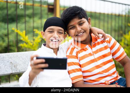 Zwei glückliche, multiethnische Kinder, die im Park Selfie auf dem Handy machen – ein Gepräch aus Freundschaft, religiöser Vielfalt und sozialer Harmonie Stockfoto