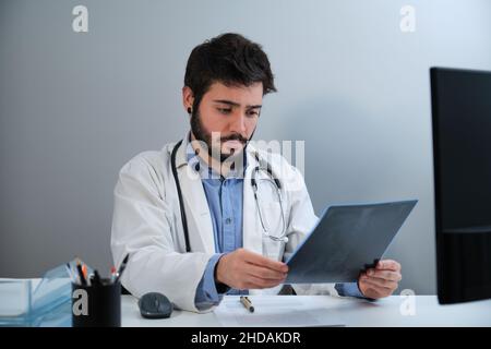 Junger Arzt, der eine Röntgenaufnahme eines Beckens und einer Wirbelsäule am Schreibtisch ansieht. Stockfoto