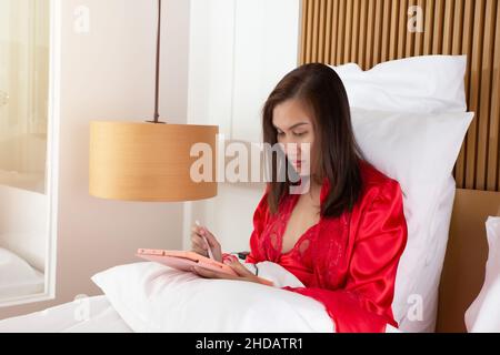 Asiatische Frau mit Tablet-pc in ihrem Bett in der Nacht. Thailänderin in einer roten Satin-Nachtwäsche mit Tablet-Computer in einem Schlafzimmer. Weibliche benutzen Tabletten auf dem Bett Stockfoto