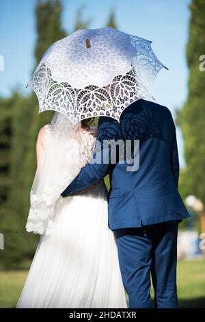 Braut und Bräutigam umarmen sich an ihrem Hochzeitstag unter einem Spitzenschirm - selektiver Fokus Stockfoto