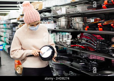 Frau untersucht die Bratpfanne in der Abteilung für Küchenutensilien im Supermarkt sorgfältig. Sie trägt eine Maske und einen rosa Strickmütze wie einen Pullover. Einkaufen in e Stockfoto