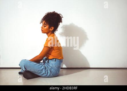 Junge ruhige nachdenkliche afroamerikanische Mädchen mit hoher Frisur in Lotushaltung auf dem Boden gegen weiße Wand mit Schatten, nachdenkliche schwarze woma sitzen Stockfoto