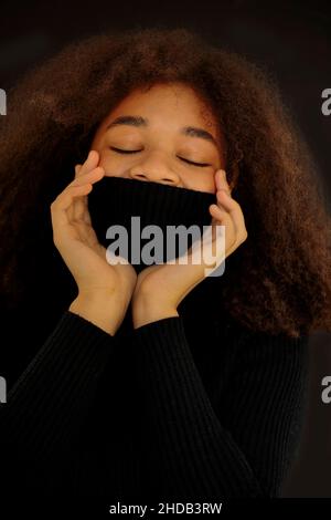 Erfreut afroamerikanische lockige Frau, die Augen schließt und Gesicht mit warmem schwarzen Pulloverkragen bedeckt, isoliert auf dunklem Studiohintergrund, lächelnd weiblich Stockfoto