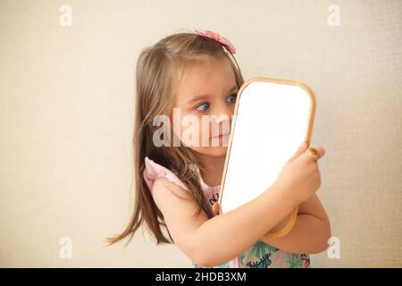 Junge neugierige kleine Mädchen halten Spiegel Blick auf ihre Reflexion während des Spielens und Freizeit zu Hause, tragen Make-up und tun es nur l Stockfoto