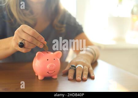 Geld sparen. Ausgeschnittene Aufnahme einer Frau, die eine Münze in ein Keramik- oder Porzellanschweinebank legt, während sie zu Hause am Tisch sitzt, selektiver Fokus. Grundlagen der Budgeti Stockfoto