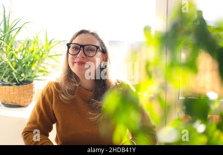 Porträt einer lächelnden Frau mittleren Alters, die eine Brille trägt und im Zimmer mit grünen Topfpflanzen sitzt, selektiver Fokus. Positive glückliche 40s weibliche Schmerzen Stockfoto