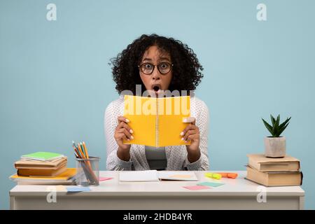 Porträt einer schockierten jungen schwarzen Frau in einer Brille, die Notizbuch liest und am Tisch auf blauem Studiohintergrund sitzt Stockfoto