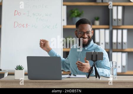 Lächelnd hübscher junger schwarzer männlicher Tutor mit Bart zeigen englische Regeln und schaut auf pc und Smartphone Stockfoto