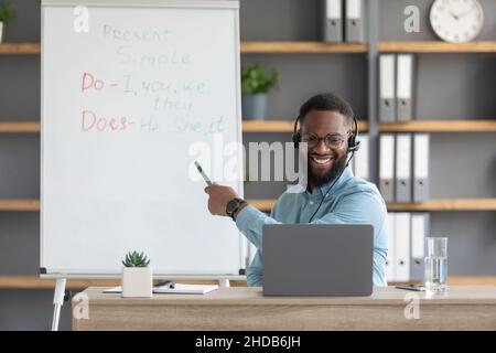 Freundlicher junger afroamerikanischer Mann Tutor zeigt Regeln des englischen an der Tafel und schaut auf den pc Stockfoto