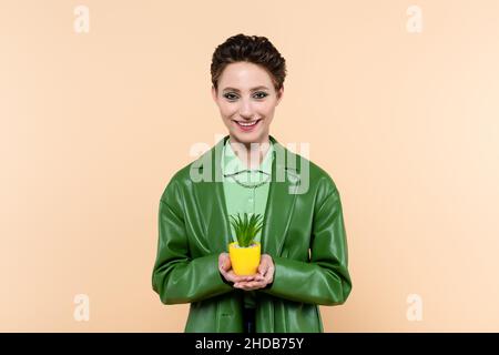 Glückliche Frau in grüner Jacke hält Pflanze in gelben Blumentopf isoliert auf beige