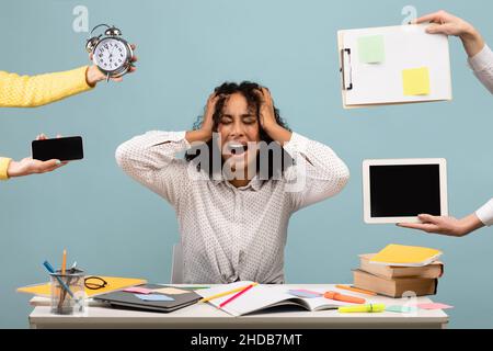 Schwarze Dame am Schreibtisch, umgeben von Händen mit Uhr und Geräten, schreiend vor Verzweiflung, an Burnout leidend Stockfoto