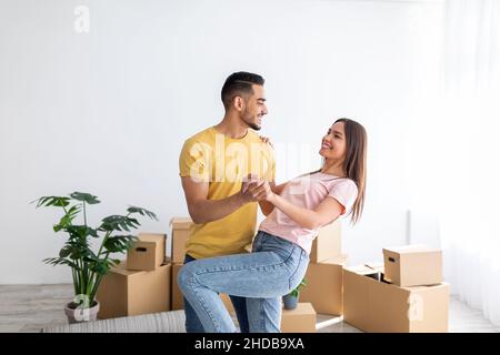 Glückliches, tausendjähriges, vielfältiges Paar, das am Umzugstag in ihrem neuen Zuhause zwischen Pappkartons tanzt, Platz zum Kopieren Stockfoto