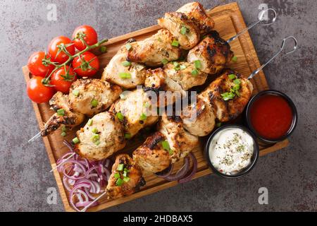 Köstliche Hühnchen-Kebabs auf Spießen mit frischem Gemüse und zwei Saucen in Nahaufnahme auf einem Holztablett auf grauem Betonboden. Horizontale Ansicht von oben Stockfoto