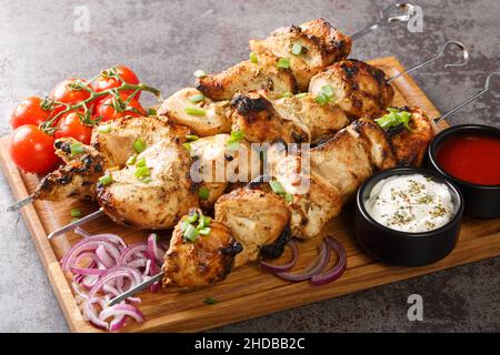 Köstliche Hühnchen-Kebabs auf Spießen mit frischem Gemüse und zwei Saucen in Nahaufnahme auf einem Holztablett auf grauem Betonboden. Horizontal Stockfoto