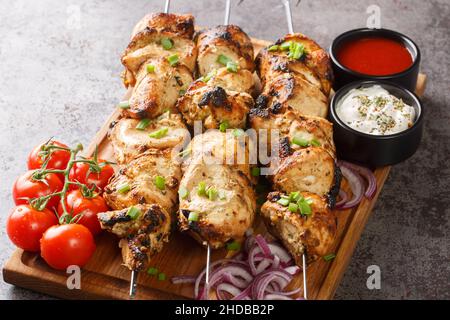 Saftig gegrillte Hähnchenspieße mit frischem Gemüse und zwei Saucen in Nahaufnahme auf einem Holztablett auf grauem Betonboden. Horizontal Stockfoto