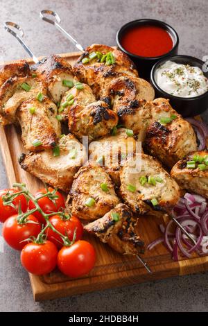 Hausgemachter Hähnchen-Kebab auf Spießen mit Gemüse und zwei Saucen in Nahaufnahme in einem Holzbrett auf grauem Beton-Hintergrund. Vertikal Stockfoto