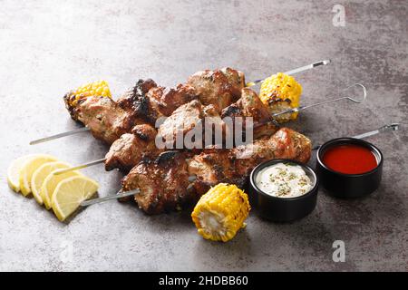 Selbstgemachte Kebabs auf Spießen mit gegrilltem Mais, Zitrone und zwei Saucen in Nahaufnahme auf grauem Beton-Hintergrund. Horizontal Stockfoto