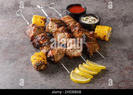 Duftendes Fleisch auf Spießen mit gegrilltem Mais und zwei Saucen in Nahaufnahme auf grauem Betongrund. Horizontal Stockfoto