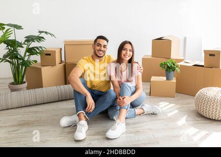In voller Länge Porträt eines liebenden multinationalen Paares, das auf dem Boden zwischen Kartons sitzt, umarmt und die Kamera anschaut Stockfoto
