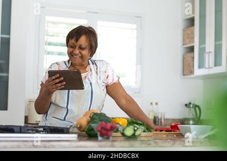 Lächelnde ältere Frau, die sich online Rezepte auf einem digitalen Tablet ansieht, während sie zu Hause in der Küche kocht Stockfoto