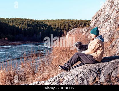Junge Frau, die auf einem Stein sitzenden Hund sitzt und den Fluss anschaut, der im Herbst oder zu Beginn des Winters mit Eis bedeckt ist, beige neutrale Farben, Erdtöne Stockfoto