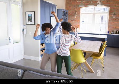 Sorglose Mutter und Tochter im Teenageralter tanzen mit aufgehobenen Armen im Wohnzimmer zu Hause Stockfoto